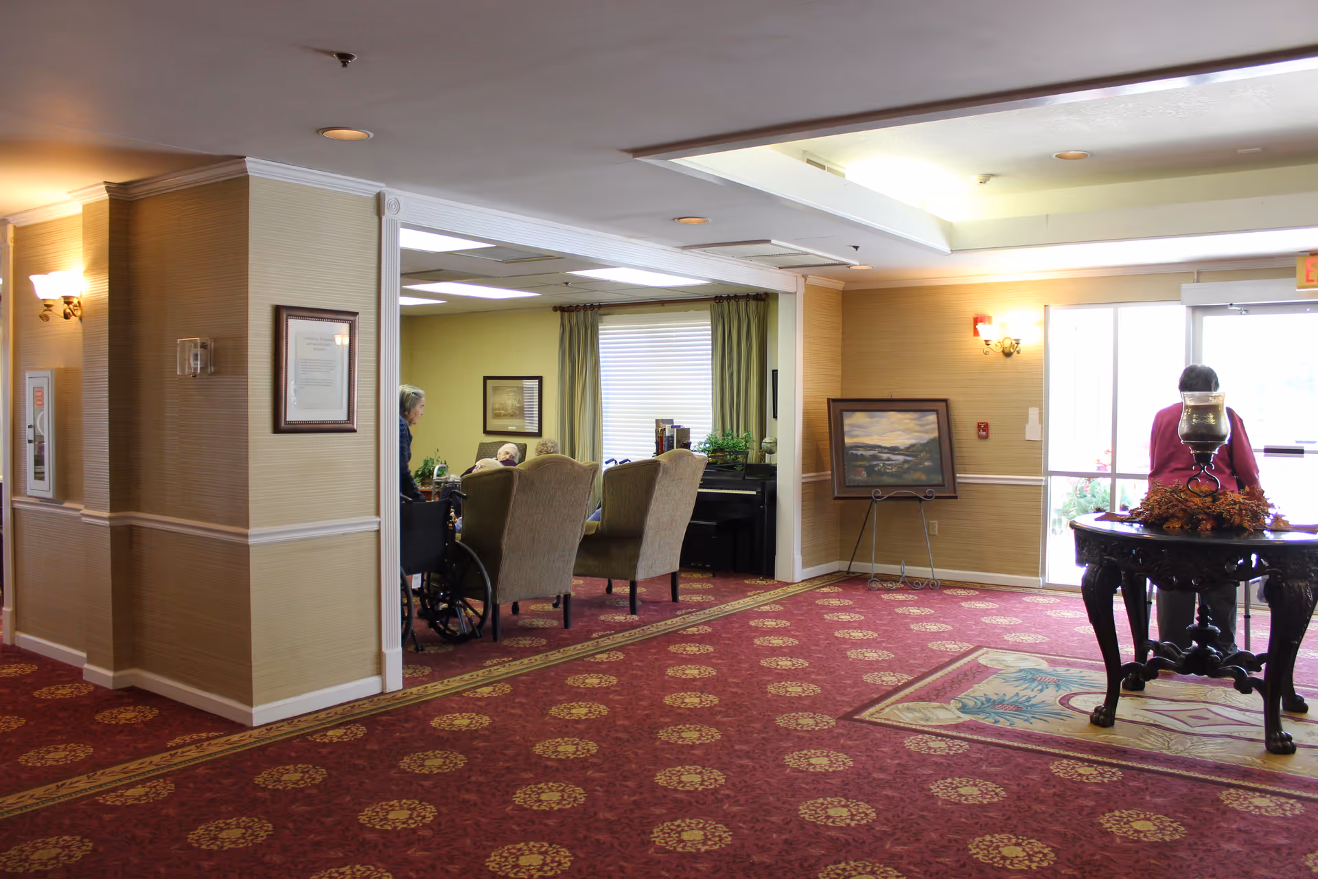Interior view of a senior living community common area with patterned red carpet, beige walls, and ceiling lights. There are two beige armchairs and a person in a wheelchair near a window with green curtains. A framed painting is displayed on an easel against the wall. A round wooden table with a decorative centerpiece is near the entrance door, where a person is standing with their back to the camera.