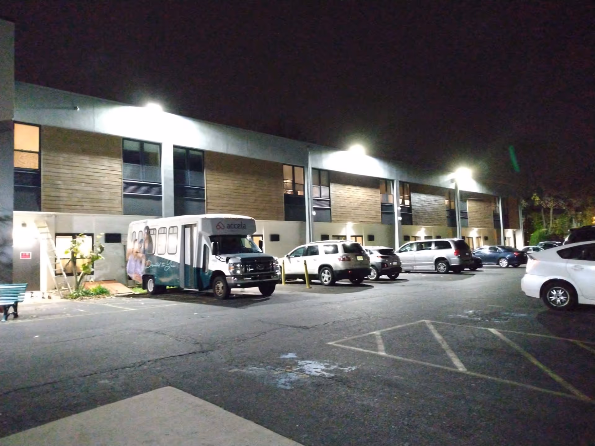 Nighttime view of the exterior of a two-story building with multiple windows and exterior lights. Several cars are parked in the parking lot in front of the building, along with a shuttle bus labeled 'accela'.