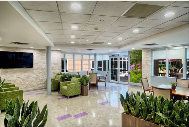 Well-lit communal lounge with green upholstered chairs, tables, potted plants, a wall-mounted TV, and glass doors/windows opening to an outdoor patio.