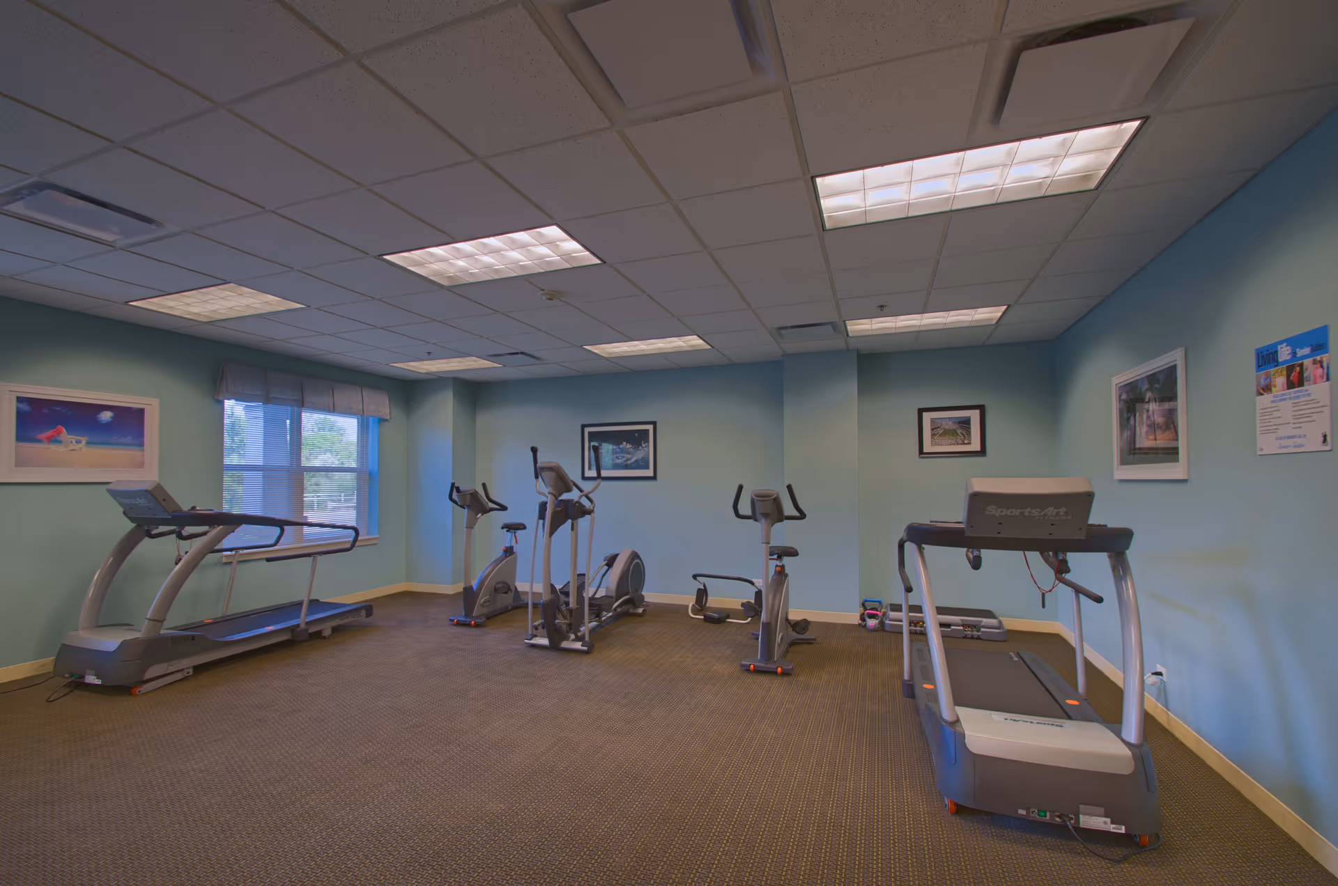 A small fitness room with light blue walls and carpeted floor containing two treadmills, two stationary bikes, and an elliptical machine. The room has fluorescent ceiling lights, a window with a valance, and framed pictures on the walls.
