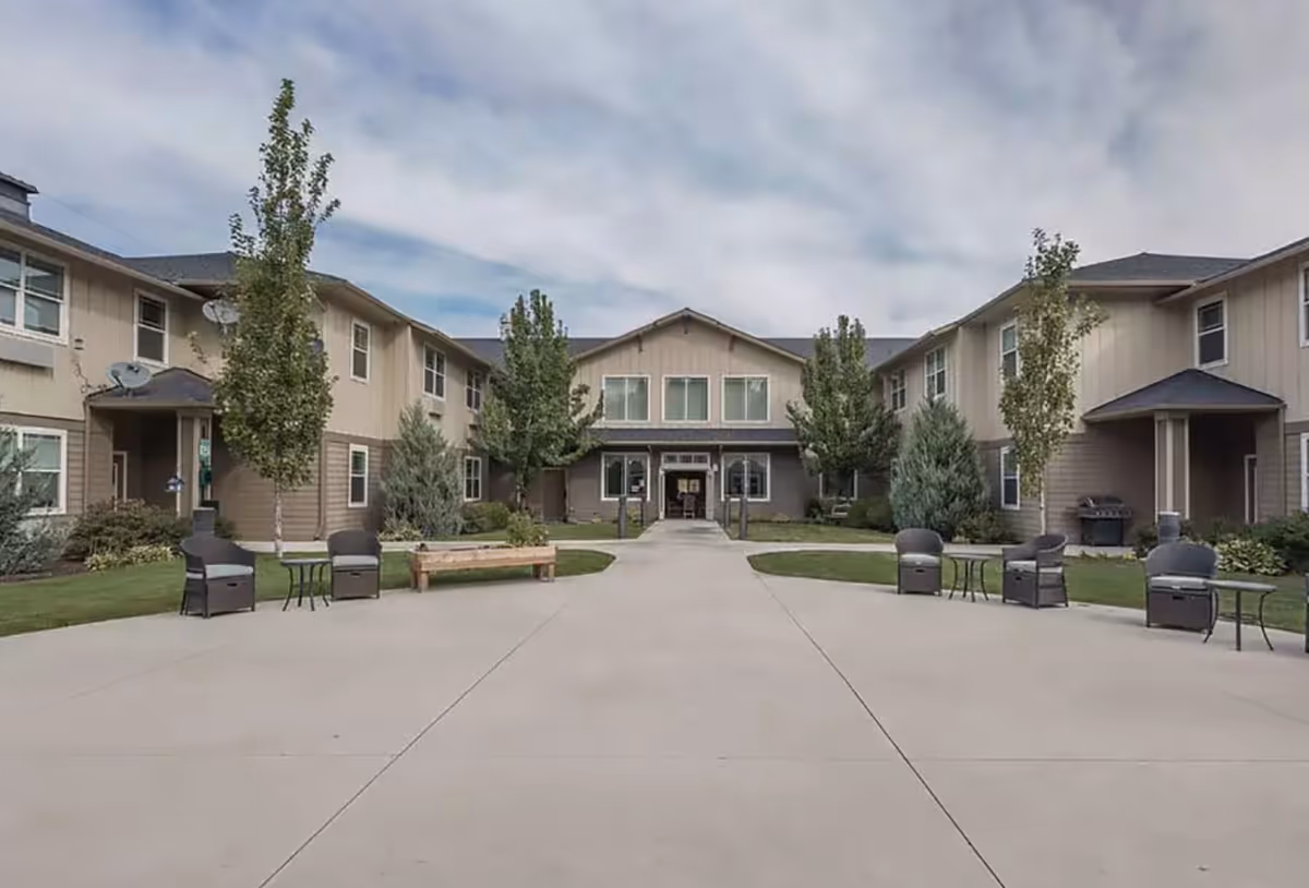 Exterior view of Regency Pullman senior living facility showing a two-story building with beige siding and multiple windows. There is a wide concrete pathway leading to the entrance, flanked by green lawns, trees, outdoor chairs, tables, and a bench. The sky is partly cloudy.