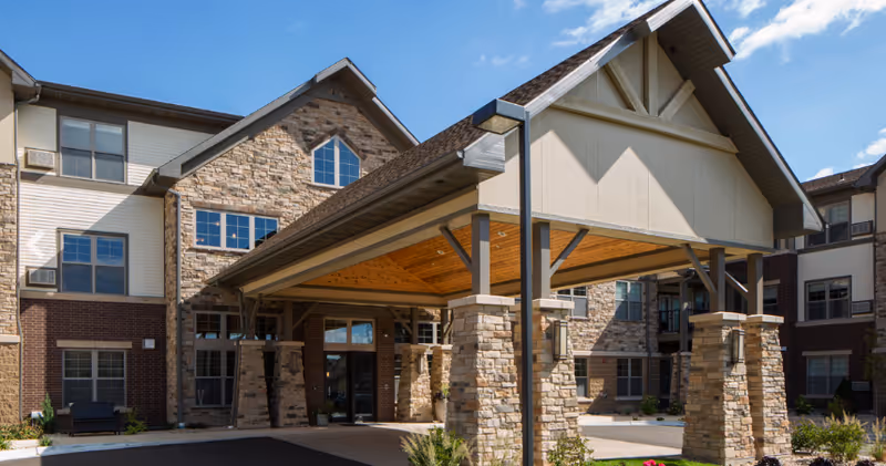 Exterior view of Noel Manor Retirement Living facility showing a large covered entrance supported by stone pillars, with a multi-story building in the background under a blue sky with some clouds.