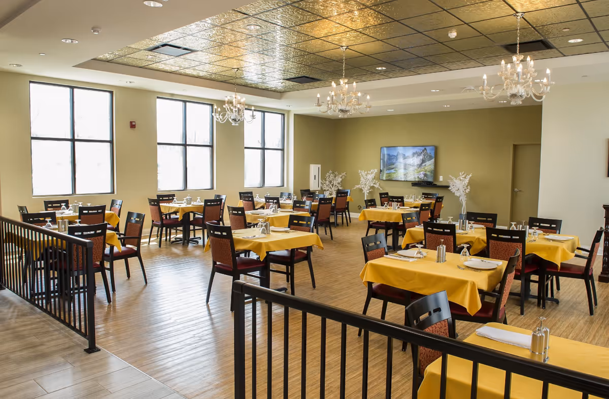 A bright dining room with multiple tables covered in yellow tablecloths, each set with plates, glasses, and silverware. The room features large windows letting in natural light, chandeliers hanging from a decorative ceiling, and a wall-mounted TV displaying a mountain landscape. The floor is wooden, and there are black chairs with patterned cushions around the tables.
