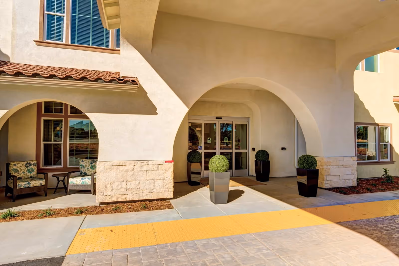 Entrance of Rose Arbor Village featuring a covered walkway with large arches, three tall planters with round green shrubs, sliding glass doors, and a small seating area with two cushioned chairs and a side table under a window.