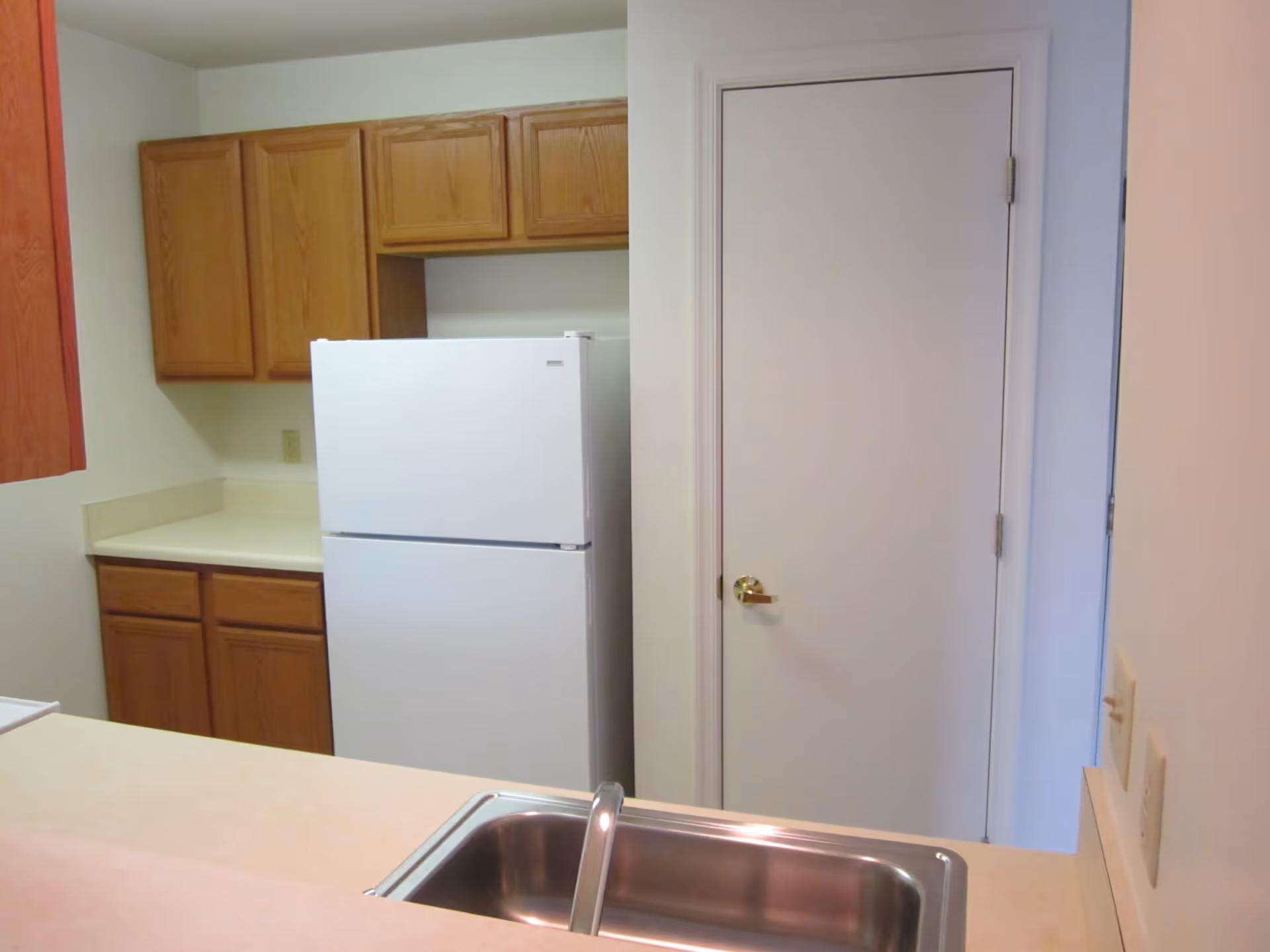 A kitchen area with wooden cabinets, a white refrigerator, a white door with a gold handle, and a stainless steel sink in the foreground.