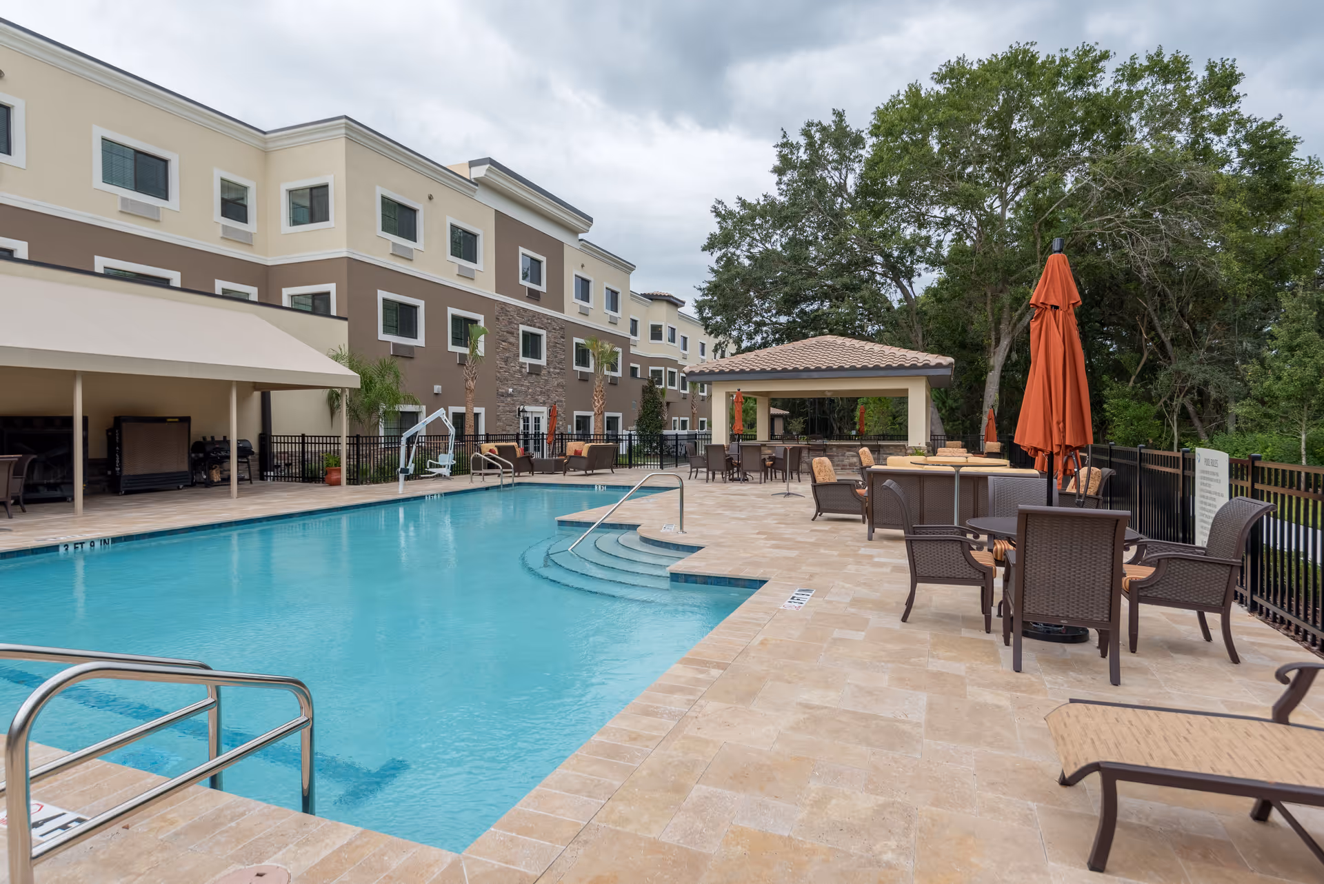 Outdoor swimming pool area at a senior living facility with lounge chairs, tables with umbrellas, and a covered seating area. The pool is surrounded by a tiled deck and a multi-story building is visible in the background along with trees.