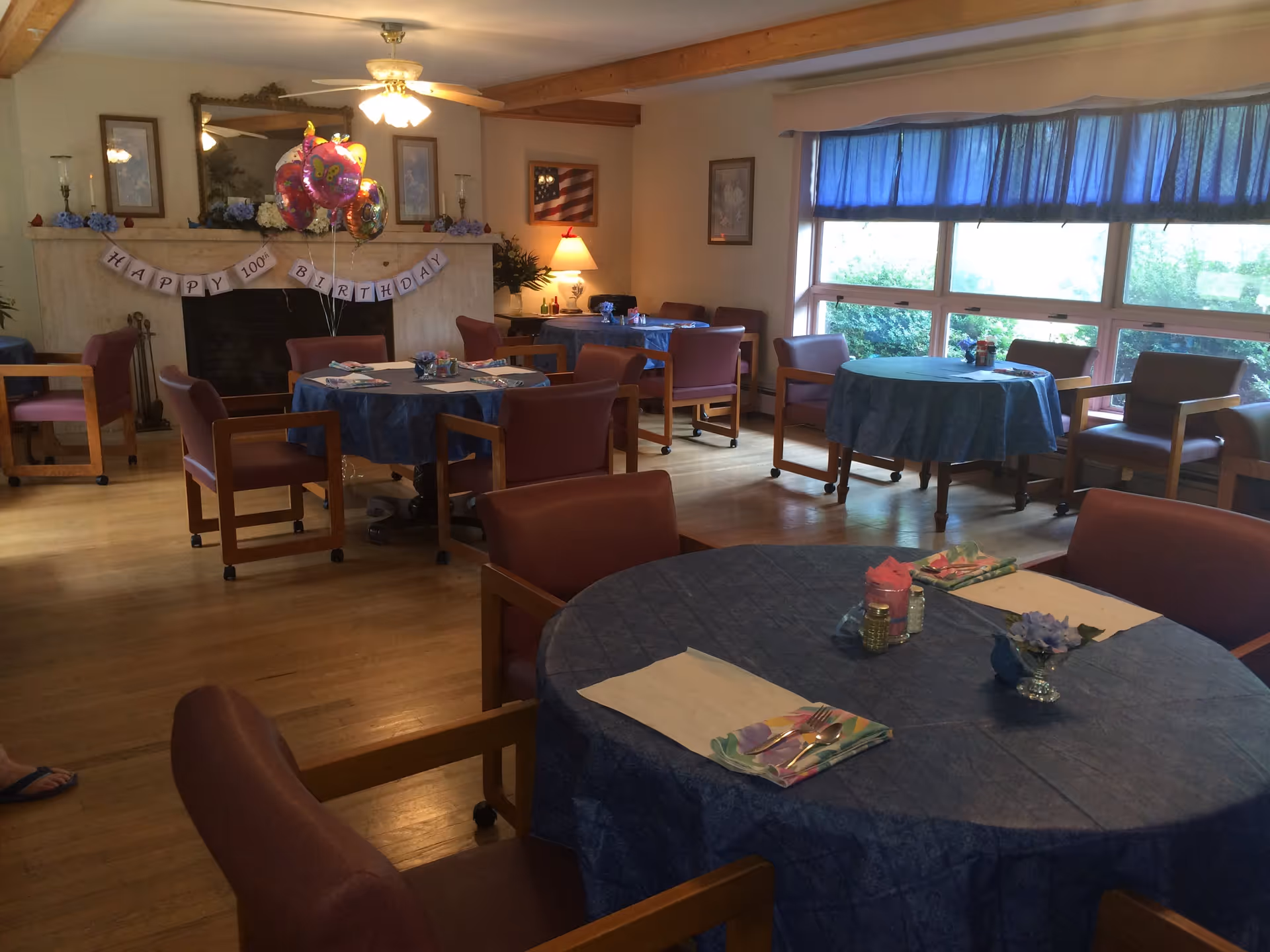 A dining room decorated for a 100th birthday celebration with round tables covered in blue tablecloths, chairs around the tables, and a banner on the fireplace that reads 'HAPPY 100th BIRTHDAY'. There are balloons near the fireplace and large windows with blue curtains letting in natural light.