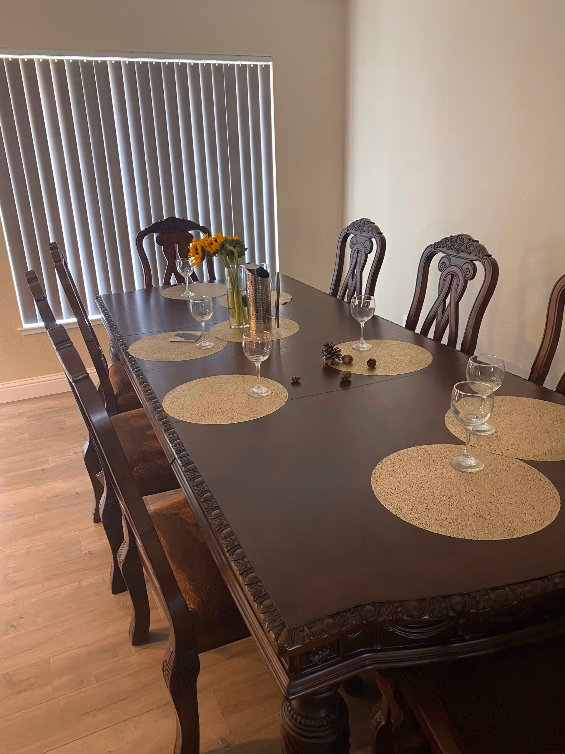 A dark wooden dining table with ornate carved edges and matching chairs arranged around it. The table is set with round beige placemats, empty wine glasses, a vase with yellow flowers, and a metal container. Vertical blinds cover the window in the background.