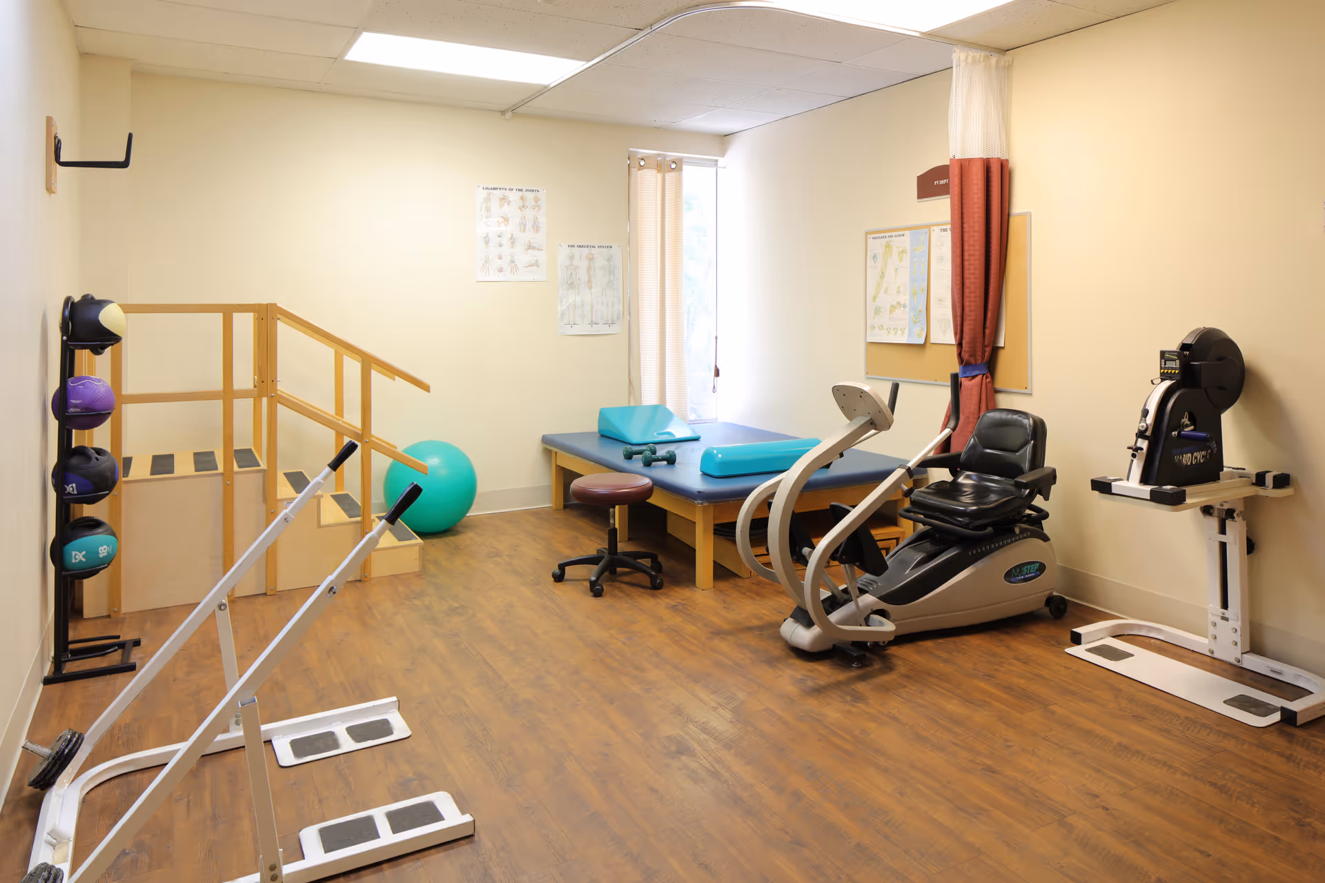 A physical therapy room with wooden flooring, featuring exercise equipment including a recumbent bike, parallel bars, a set of stairs with handrails, exercise balls, and a padded therapy table with cushions. Anatomical charts are displayed on the walls and a window with curtains lets in natural light.