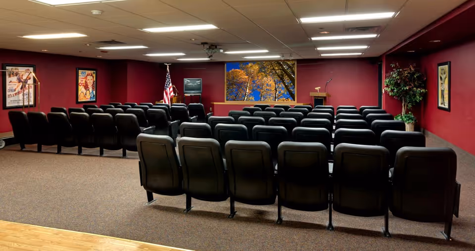 A small auditorium or presentation room with rows of black chairs facing a podium and a TV on a stand. The walls are painted dark red and decorated with framed posters. An American flag stands near the podium, and a large window shows trees with autumn leaves outside. A potted plant is placed in the corner of the room.