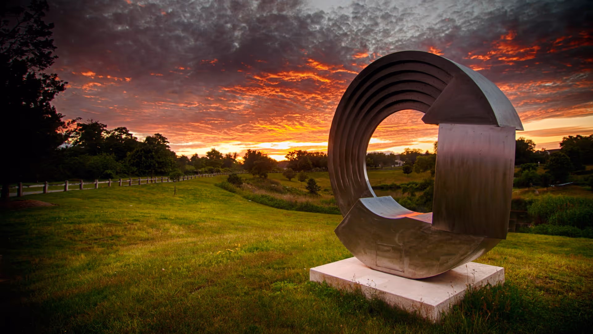A large modern metal sculpture shaped like a circular wave is displayed on a concrete base in a grassy outdoor area during a vibrant sunset with dramatic clouds and orange sky.