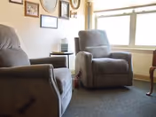 A cozy room with two gray recliner chairs positioned near a window with natural light. The wall behind the chairs is decorated with several framed pictures and a round mirror. A small side table with a tissue box is placed between the chairs.