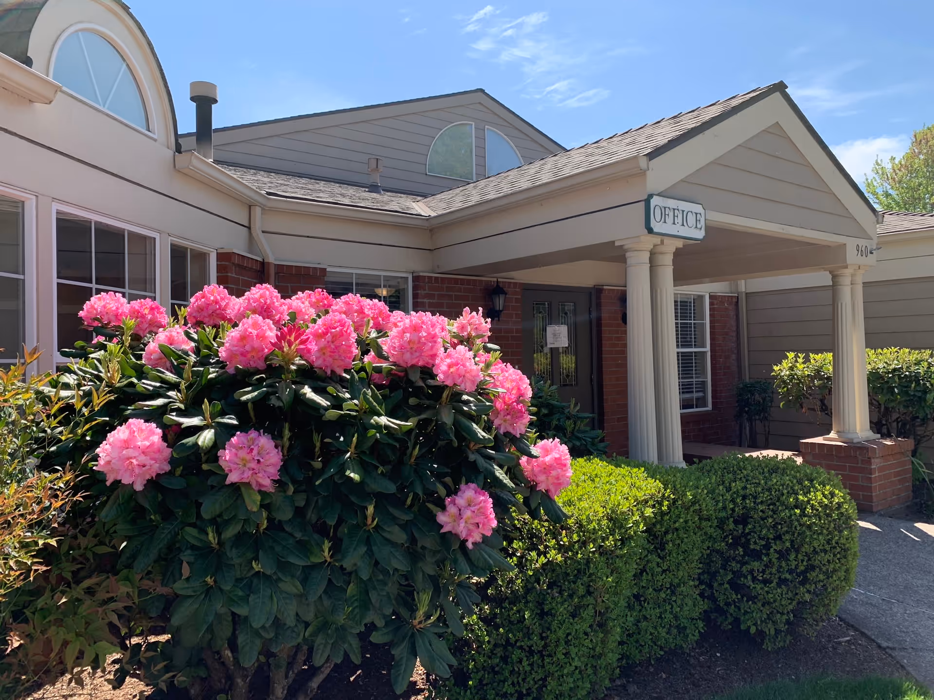 Exterior view of the Faye Wright Senior Living facility office entrance with a covered porch supported by columns, surrounded by green bushes and a large flowering shrub with pink blossoms under a clear blue sky.