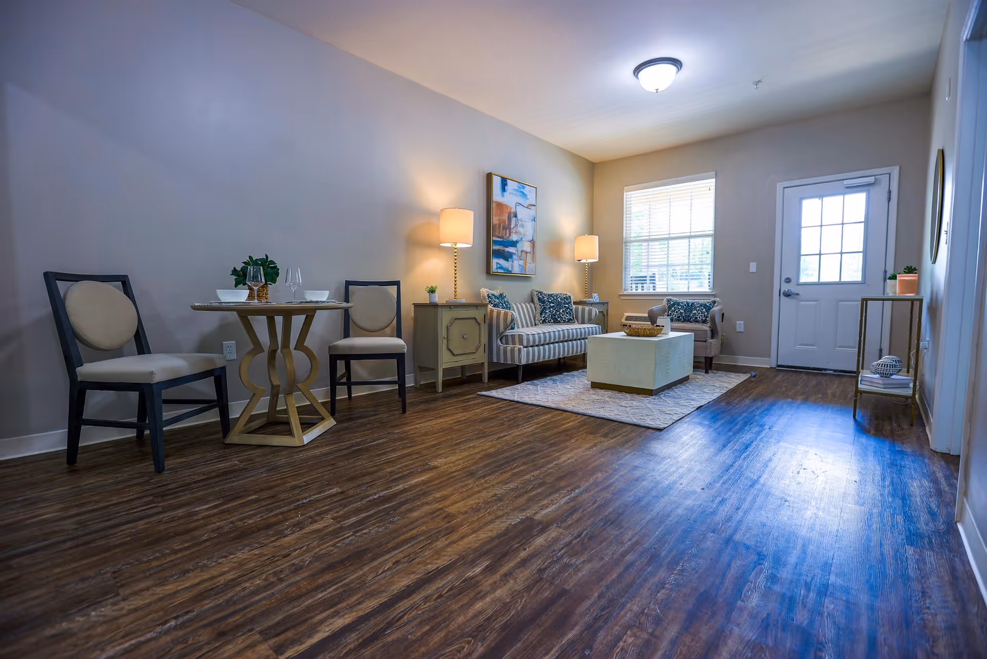 A bright living room with wooden flooring featuring a small round dining table with two chairs on the left, a striped sofa and an armchair with patterned cushions, two side tables with lamps, a coffee table on a rug, a window with blinds, and a door with glass panels on the right.