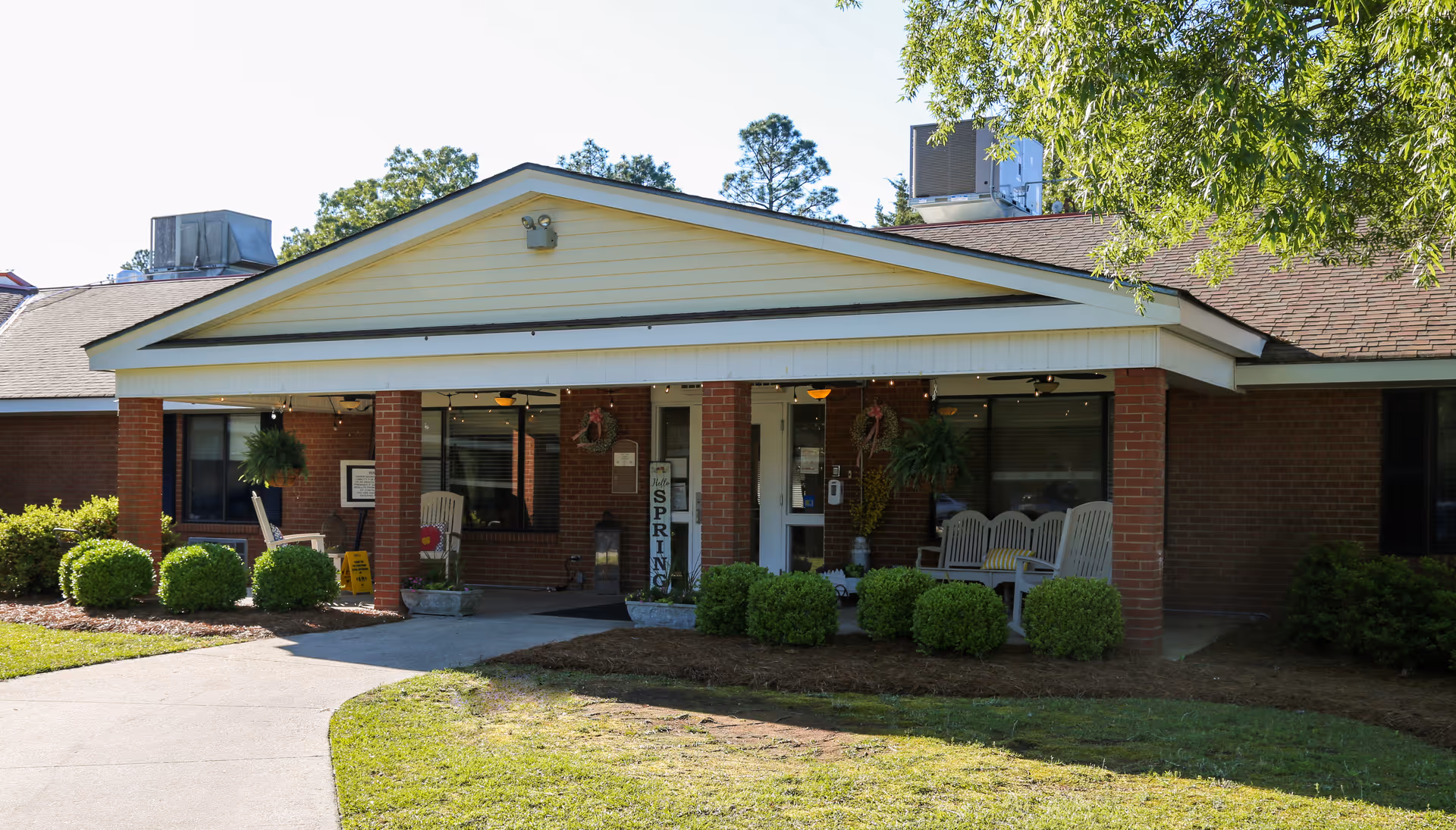 Front exterior view of a single-story brick building with a covered entrance supported by brick columns. There are white rocking chairs on the porch, green bushes lining the walkway, and trees in the background under a clear sky.