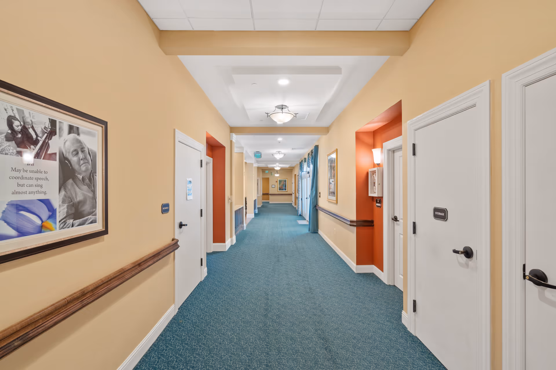 A well-lit hallway in a senior living facility with beige walls, teal carpet, and white doors on both sides. The hallway features framed artwork, handrails, ceiling lights, and an exit sign in the distance.