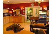 Interior view of a dining area in a senior living facility with a buffet counter on the left side, a doorway leading to a kitchen area in the center, and a seating area with tables and chairs in the foreground. The walls are painted red, and there is a chandelier hanging from the ceiling.