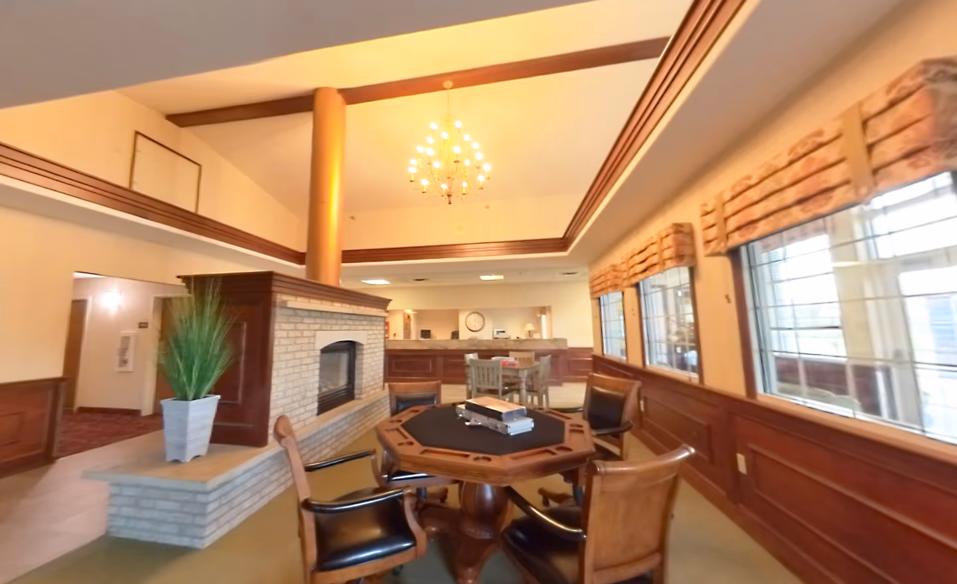 Interior view of a senior living facility common area with a central fireplace, a round table with four chairs, large windows with valances, and a chandelier hanging from a high ceiling.