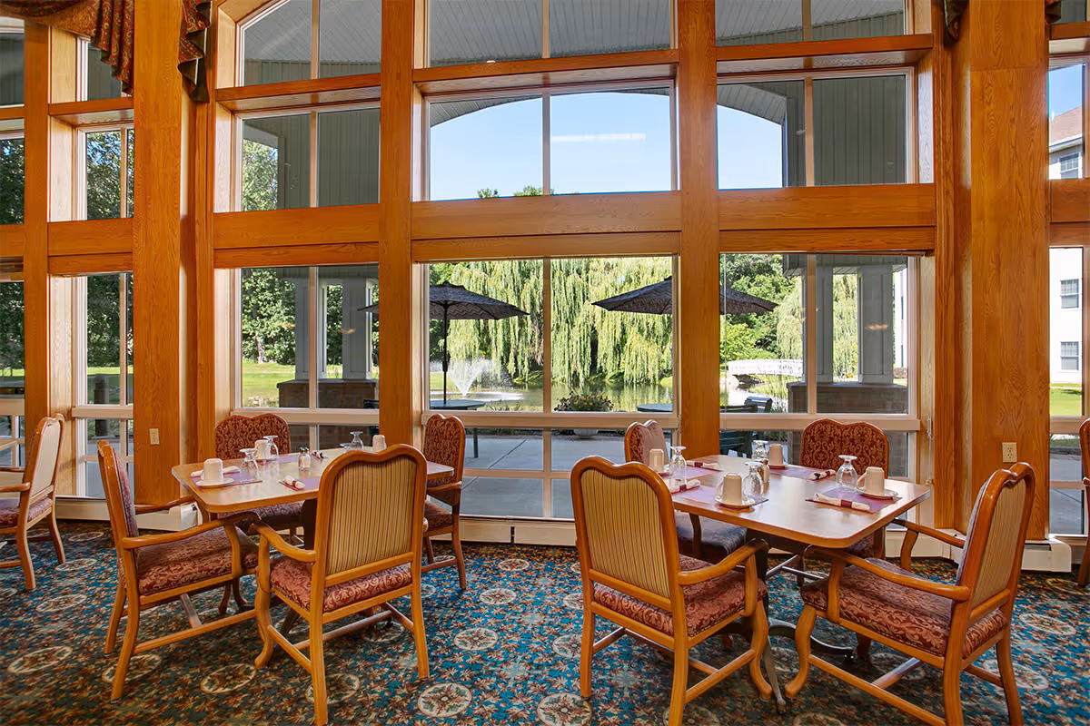 Dining area with wooden framed large windows overlooking a patio with umbrellas, a pond with a fountain, and lush greenery including a weeping willow tree. The room has carpeted flooring with a floral pattern and wooden chairs with upholstered seats and backs around wooden tables set with cups, glasses, and napkins.