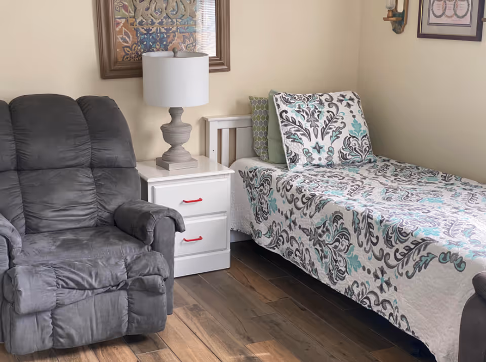 A small bedroom with a single bed in patterned bedding, a white nightstand and lamp, and a gray recliner chair.
