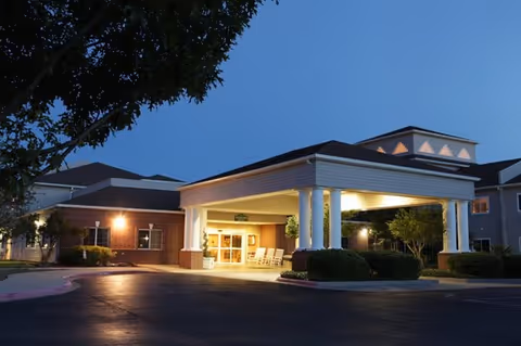 Exterior view of Lyndale San Angelo Senior Living facility at dusk, showing the entrance with a covered drop-off area supported by white columns, illuminated by warm lights. The building has brick and siding walls with a tree and bushes around the entrance.