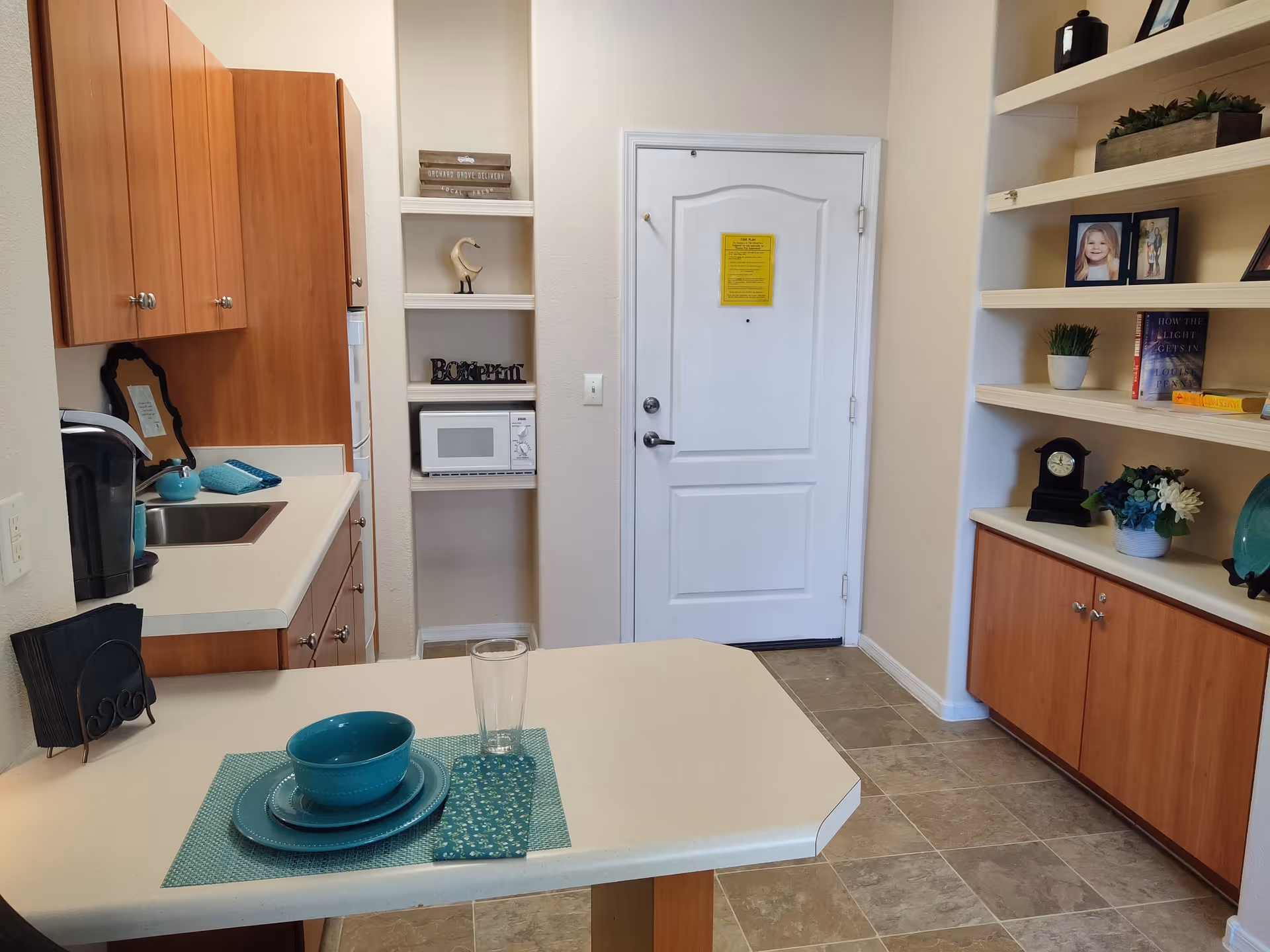 A small kitchen area with wooden cabinets, a white countertop with a teal place setting including a bowl, plate, and glass. There is a microwave on a built-in shelf next to a white door. On the right side, there are built-in shelves with framed photos, books, plants, and decorative items.