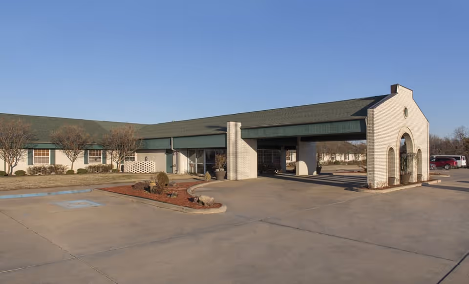 Front exterior of a single-story nursing facility with a covered porte-cochère, landscaping, and a parking lot under a clear blue sky.