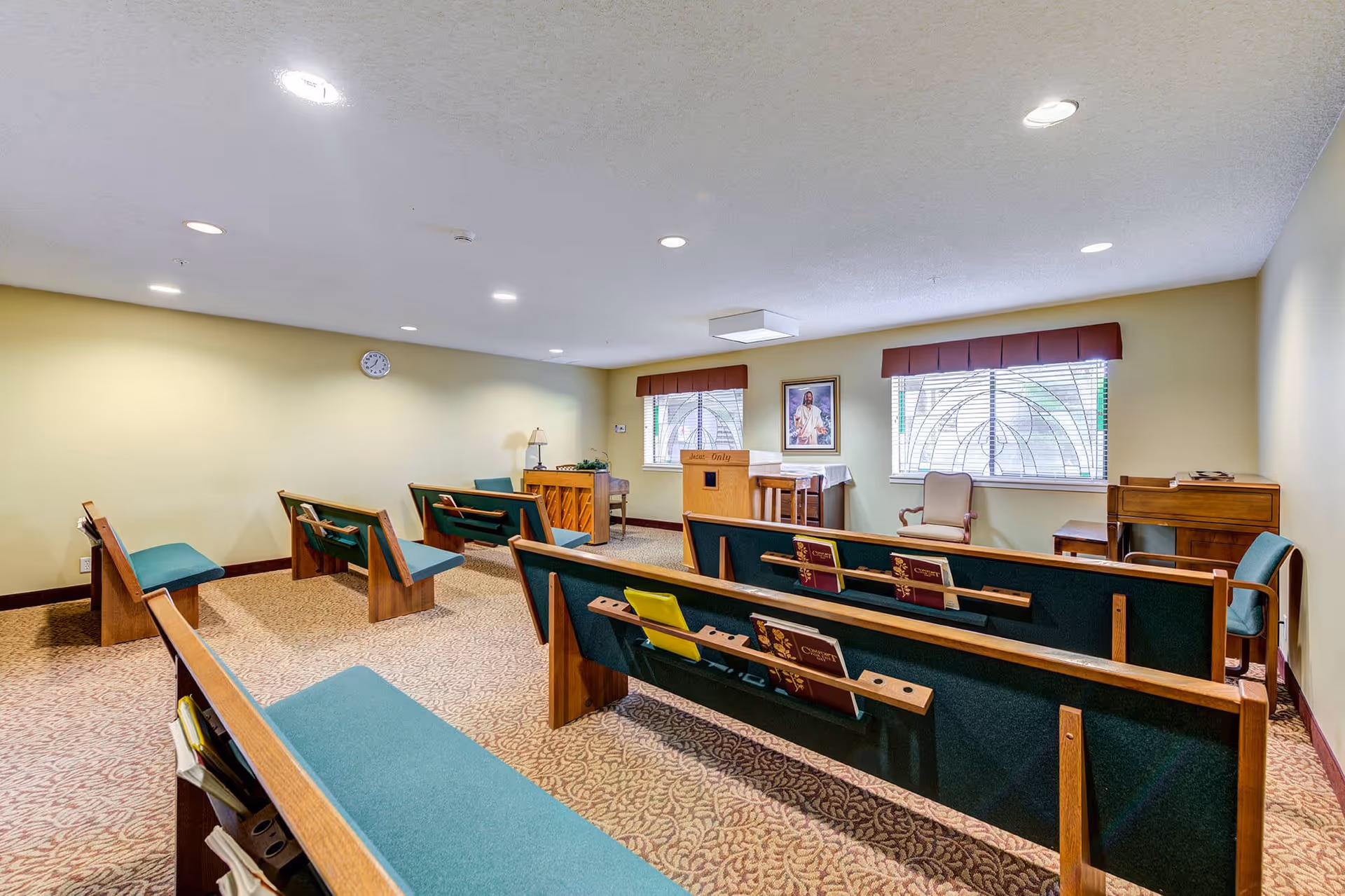 Interior of a small chapel or prayer room with wooden pews featuring green cushions, a podium, a piano, and a framed religious picture on the wall between two windows with decorative glass. The room has beige walls and carpeted floor.