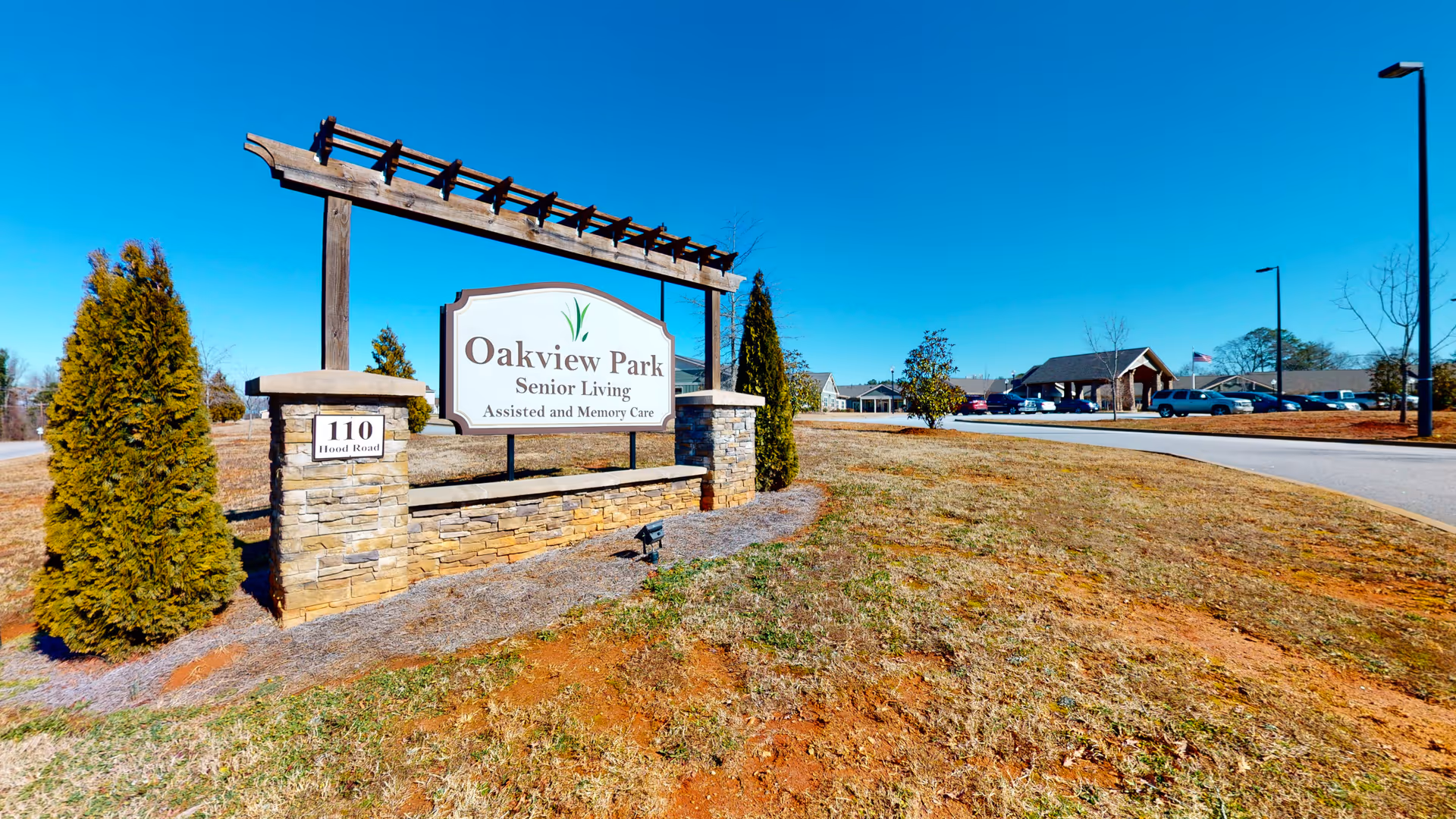 Outdoor view of the entrance sign for Oakview Park Senior Living, Assisted and Memory Care, located at 110 Hood Road. The sign is mounted on a stone base with wooden beams above it. In the background, there is a driveway leading to buildings and parked cars under a clear blue sky.