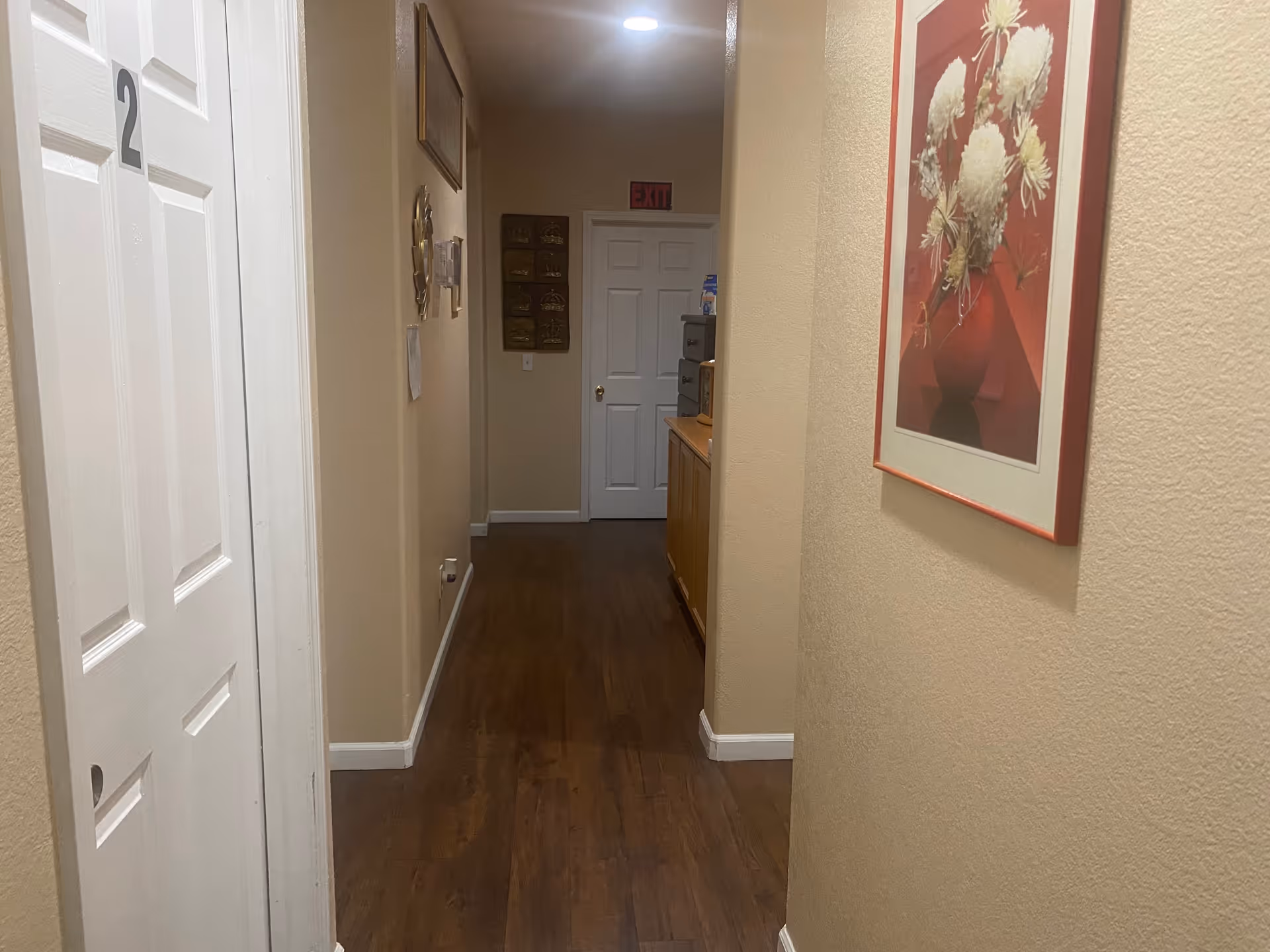 Interior hallway in a care home with wood flooring, a door marked '2', framed artwork on the wall, and an exit sign at the far end.