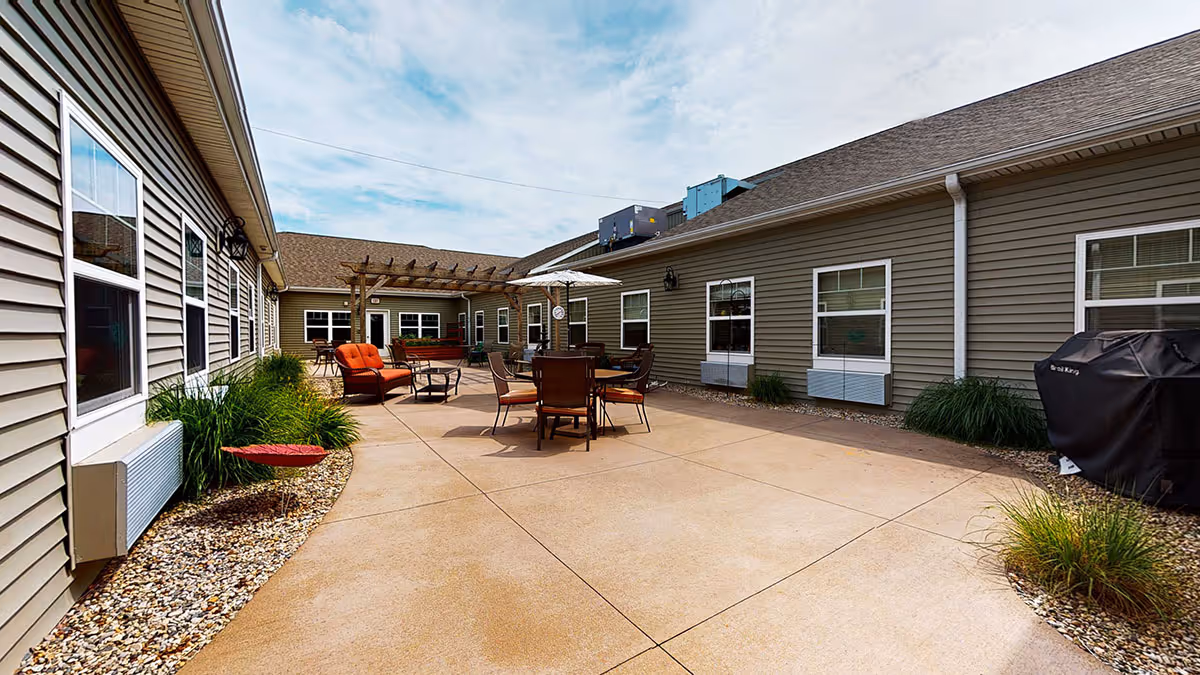 Outdoor patio area at Vicinia Gardens Luxury Retirement Living - Intermediate Care with seating arrangements including chairs, tables, an umbrella, and a pergola. The patio is surrounded by beige siding buildings with multiple windows and some greenery along the edges.