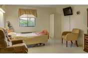 A simple, clean bedroom in a healthcare facility with a single bed covered in a yellow bedspread, a window with a valance, a beige armchair, a small wall-mounted TV, a white wardrobe, and a wooden dresser.