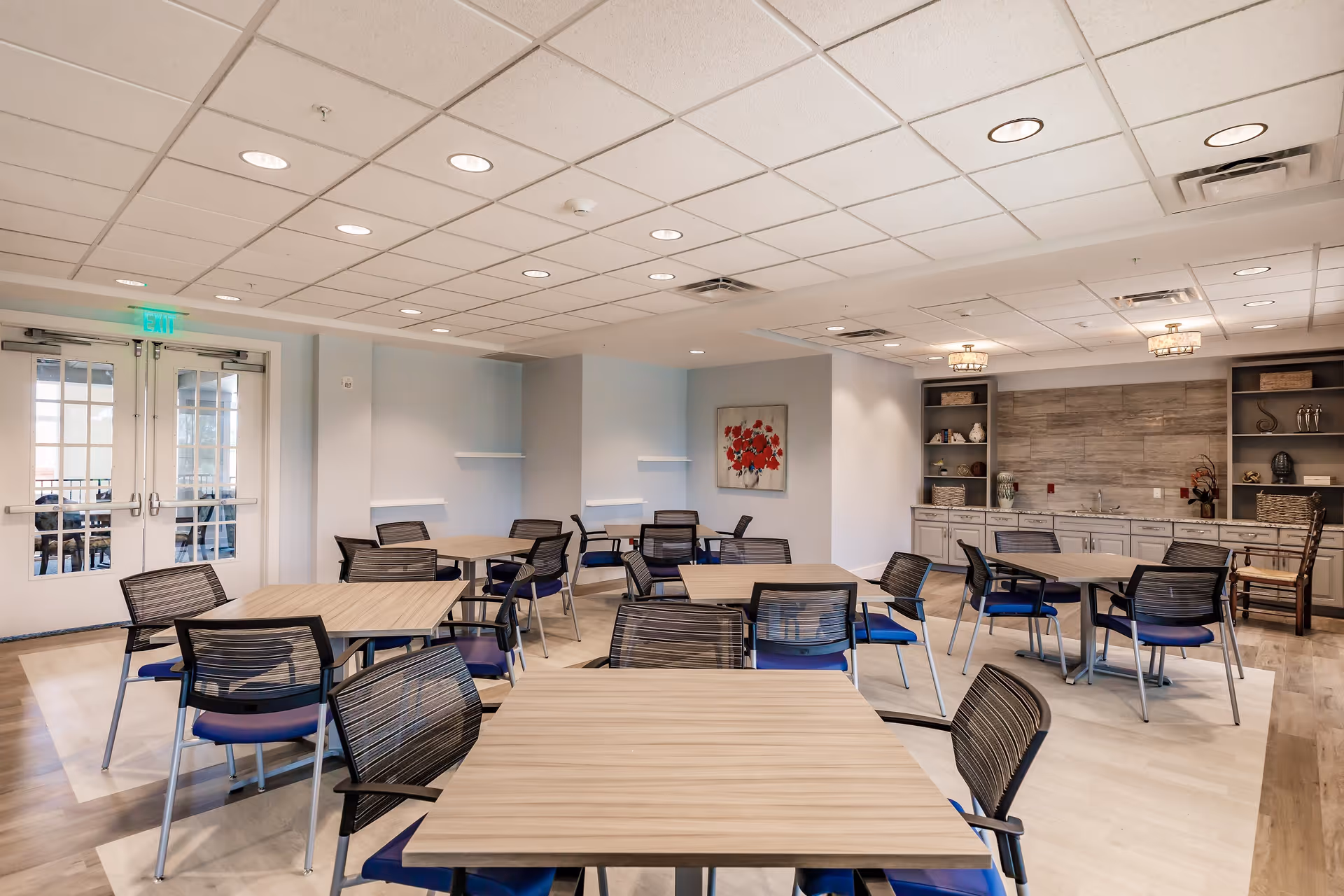 A bright and modern dining room with several square wooden tables and black mesh chairs with blue cushions. The room has a drop ceiling with recessed lighting, light-colored walls, and a decorative painting of red flowers on one wall. There is a built-in cabinet area with shelves and decorative items, and double glass doors leading to another room.