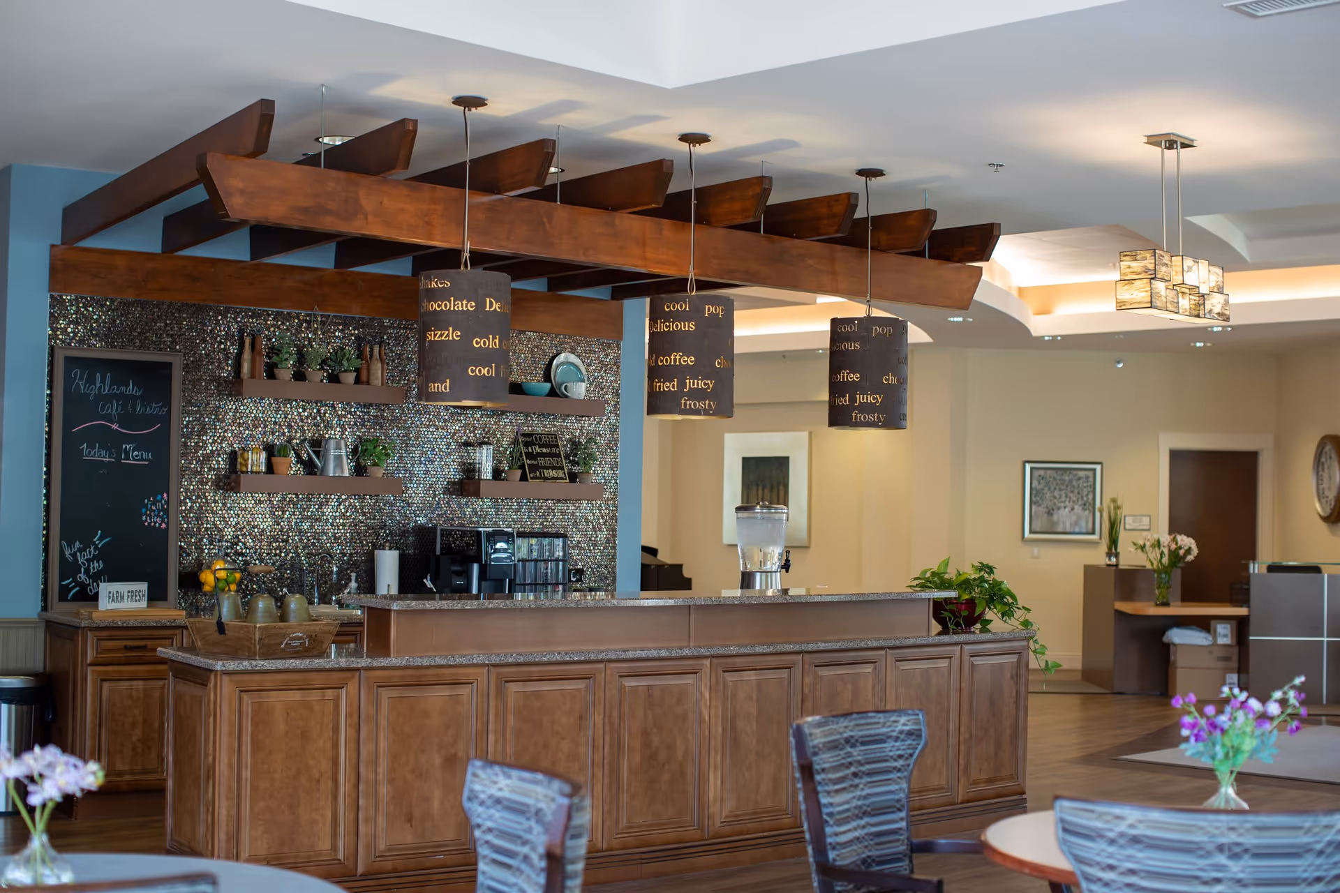 Interior view of a senior living facility's café area with a wooden counter, hanging pendant lights, shelves with plants and decor, a coffee machine, and a water dispenser. There are tables and chairs in the foreground with flower vases, and the background shows a hallway and framed artwork on the walls.