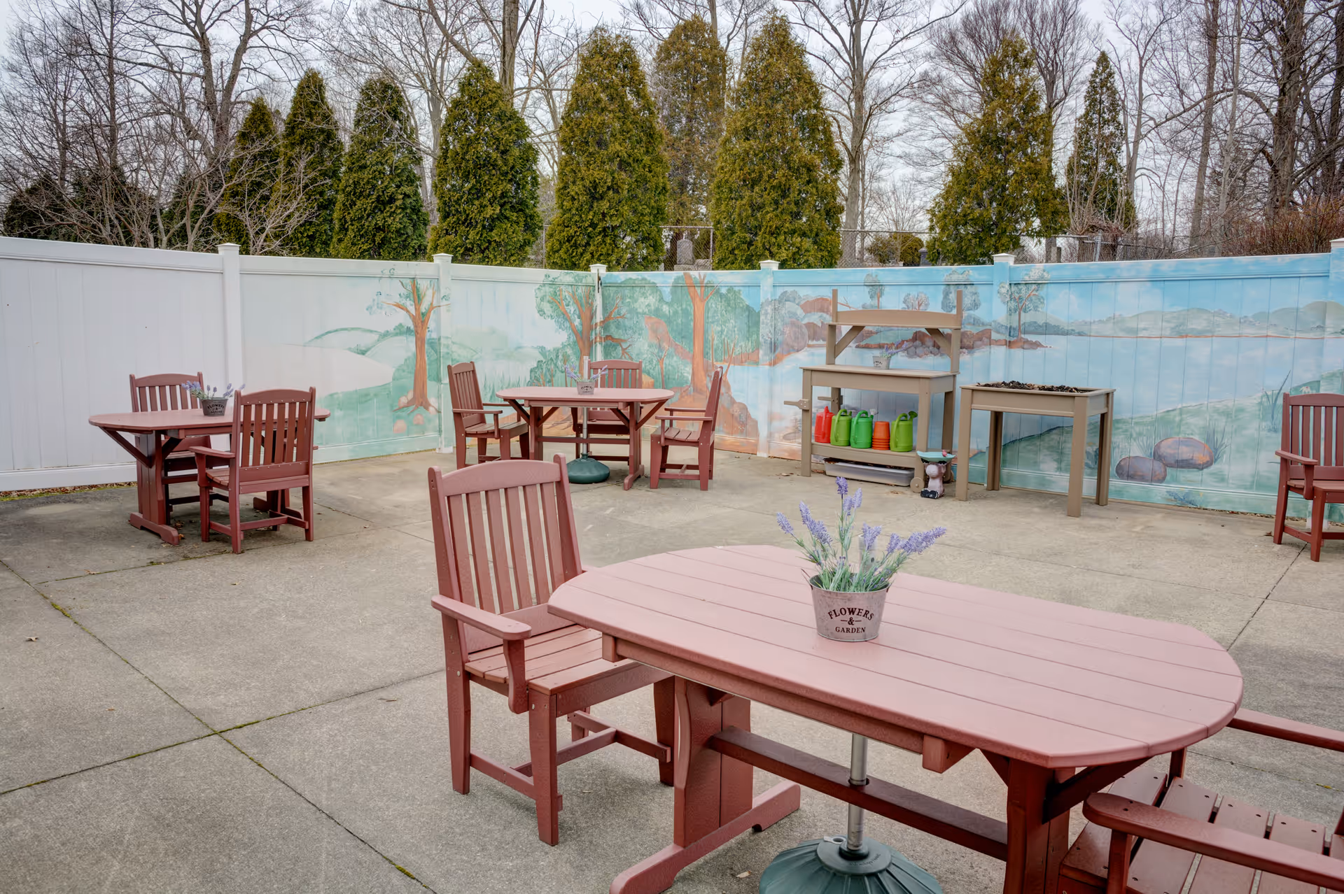 Outdoor patio area with multiple wooden tables and chairs arranged on a concrete surface. A white fence with a painted mural of trees and landscape surrounds the area. Gardening tools and colorful watering cans are placed on a shelf against the fence. Tall evergreen trees and leafless deciduous trees are visible beyond the fence.