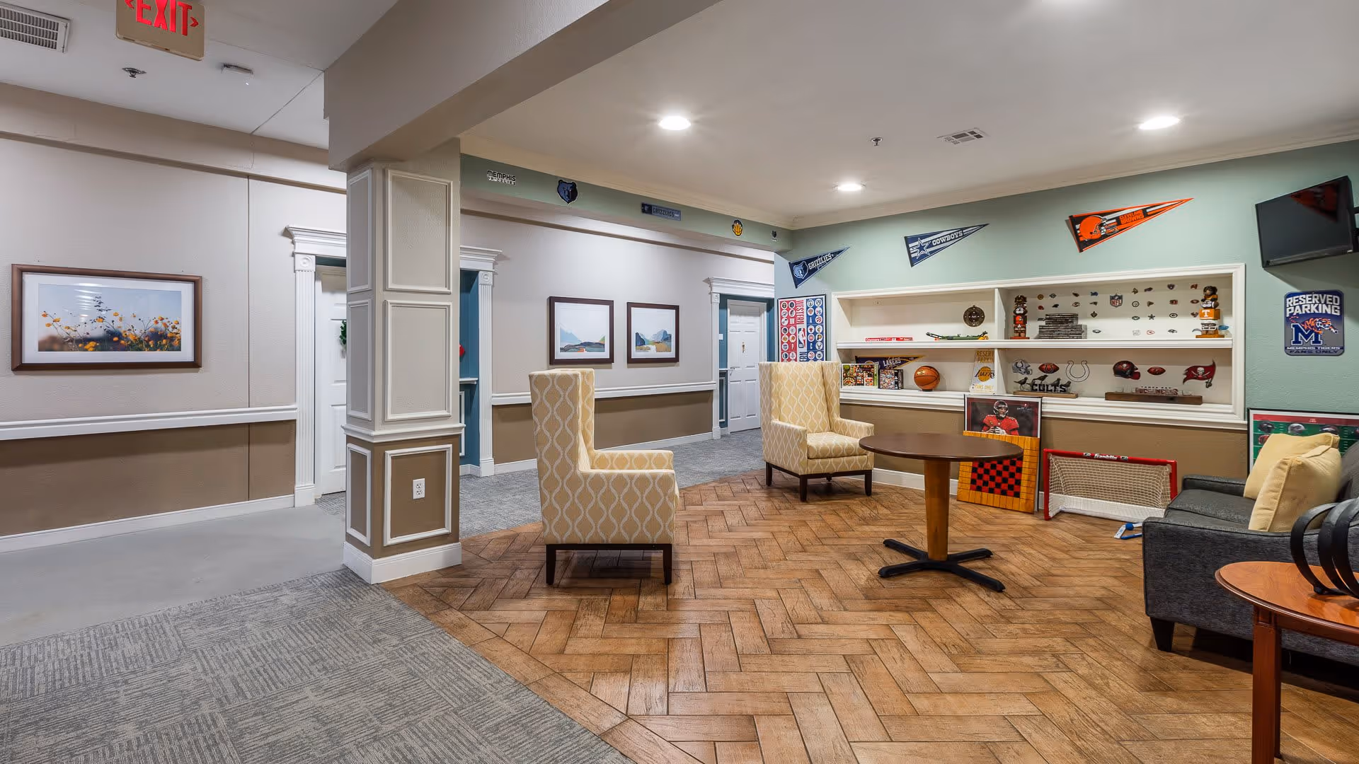 A cozy common area in Hearthstone Assisted Living featuring two patterned armchairs, a round wooden table, and a gray sofa with cushions. The walls are decorated with sports memorabilia including pennants, helmets, and various sports items displayed on built-in shelves. The floor transitions from carpet to wood in a herringbone pattern. There are framed pictures on the walls and an exit sign above a doorway.