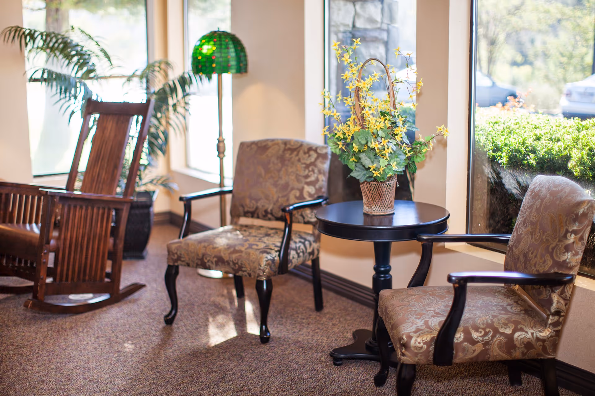 A cozy sitting area in a care center with two patterned armchairs and a round wooden table between them holding a basket of yellow flowers. A wooden rocking chair and a green stained-glass floor lamp are near large windows letting in natural light. Outside the windows, greenery and parked cars are visible.