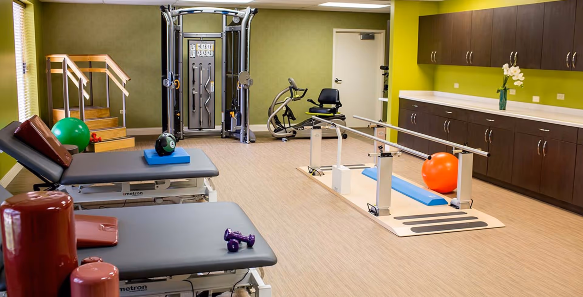 Physical therapy/exercise room with treatment tables, parallel bars, various exercise equipment, and cabinets.