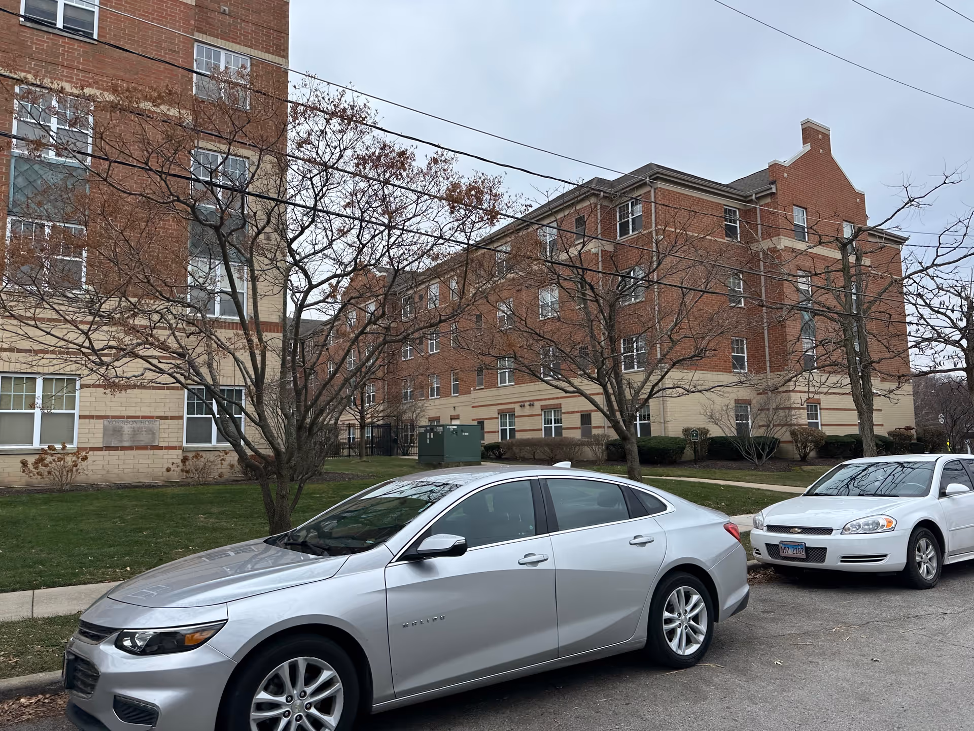 Front view of a multi-story red-brick residential building with leafless trees and two parked cars along the street.