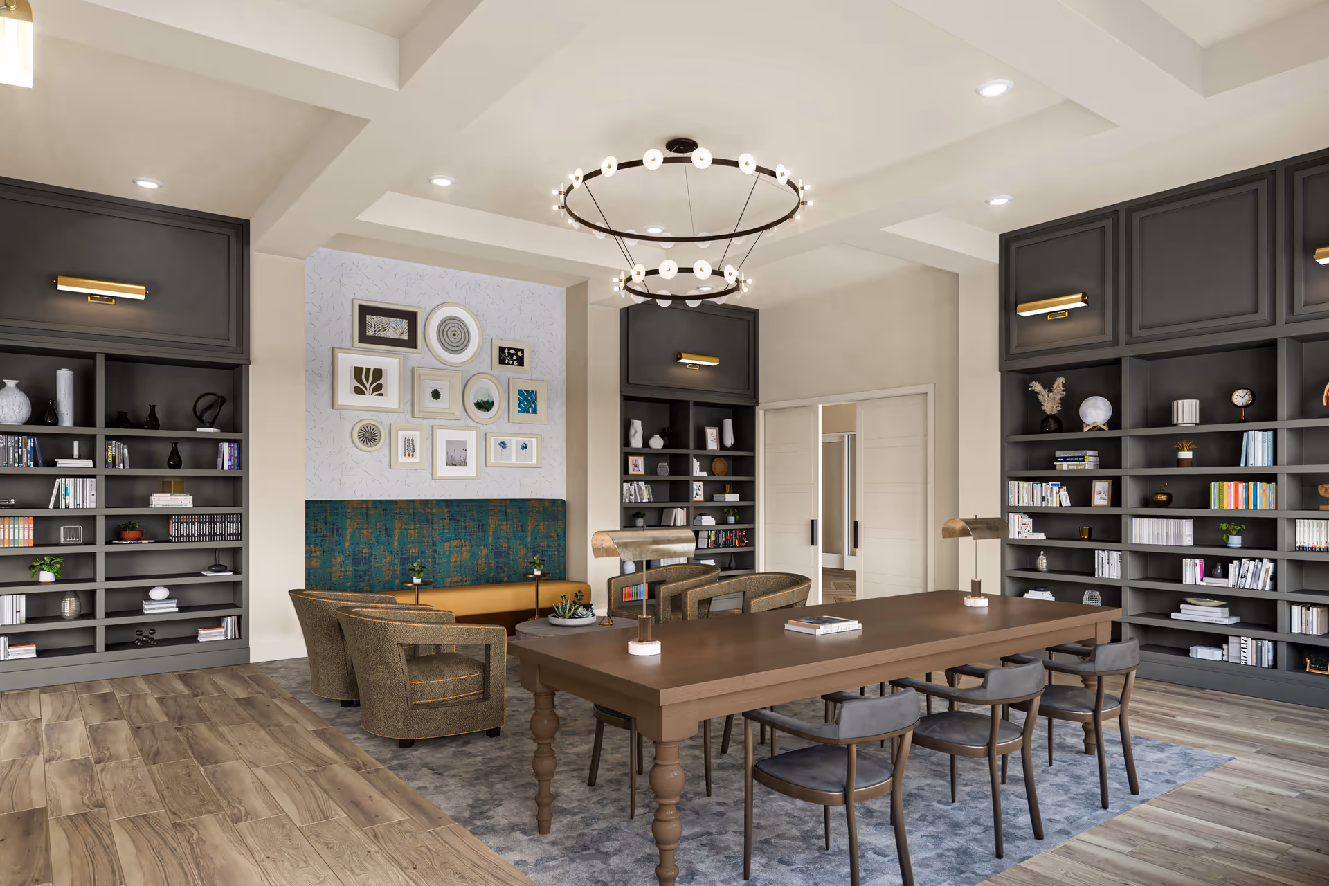 A modern and cozy interior room featuring a large wooden table with six chairs, two desk lamps, and books on the table. The room has built-in dark gray bookshelves filled with books and decorative items on both sides. There is a seating area with four upholstered chairs and a bench with a teal and gold patterned backrest against a wall adorned with framed artwork. The ceiling has recessed lighting and a large circular chandelier with multiple small lights. The floor is a combination of wood and a gray area rug.