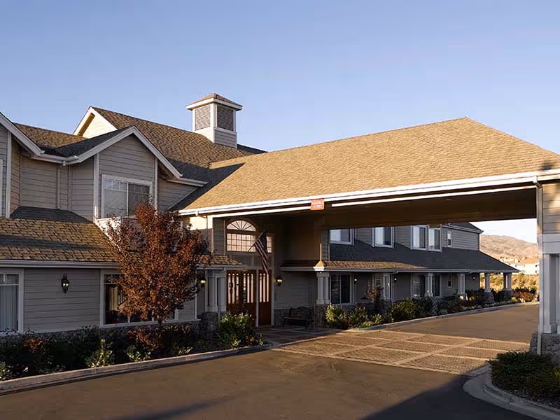 Exterior view of a senior living facility building with a covered entrance driveway, beige siding, multiple windows, and a small tree and shrubs in front.