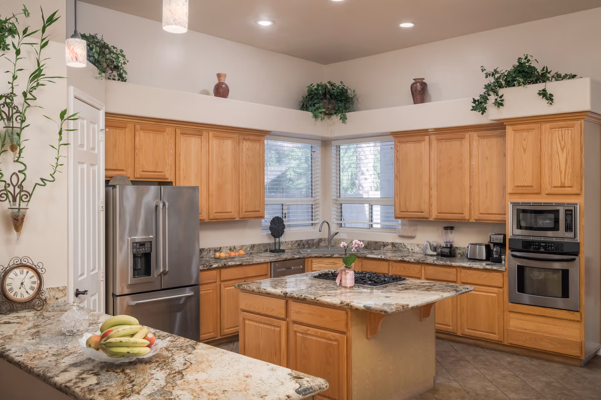 A spacious kitchen with wooden cabinets and granite countertops. The kitchen features a stainless steel refrigerator, built-in microwave and oven, a gas stove on a central island, and various small appliances like a toaster and blender. There are two windows with blinds letting in natural light, and decorative plants and vases placed above the cabinets. A bowl of bananas and other fruits is on the counter.