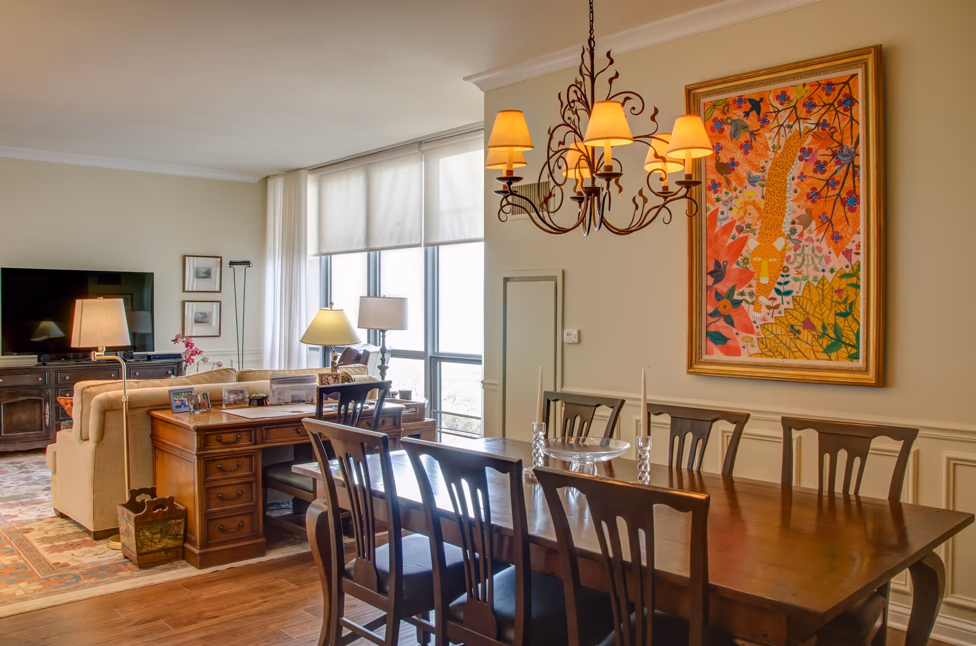 A well-lit dining and living room area with a wooden dining table and six chairs, a decorative chandelier with six lampshades hanging above the table, and a colorful framed painting on the wall. The living room features a beige sofa, a wooden desk with a chair, two table lamps, a flat-screen TV on a dark wooden cabinet, and large windows with white curtains allowing natural light to fill the space.
