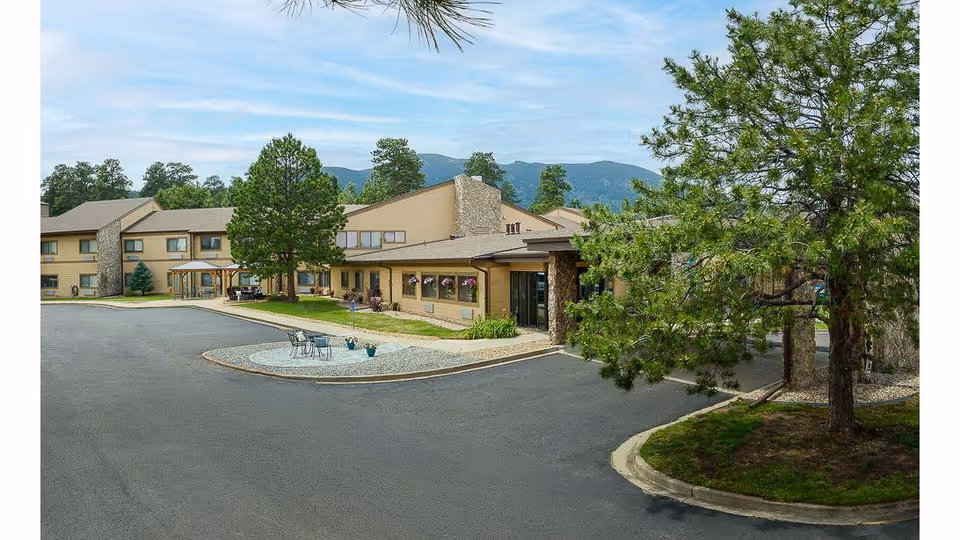 Exterior view of Life Care Center of Evergreen, a two-story building with beige siding and stone accents, surrounded by trees and mountains in the background. There is a paved driveway and a small seating area with chairs and tables near the entrance.