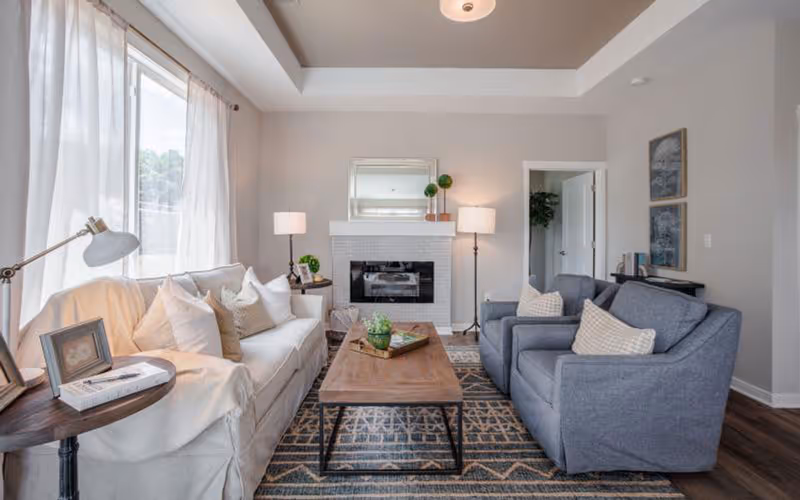Bright furnished living room with a sofa, two armchairs, a wooden coffee table, and a fireplace beneath a mirror.