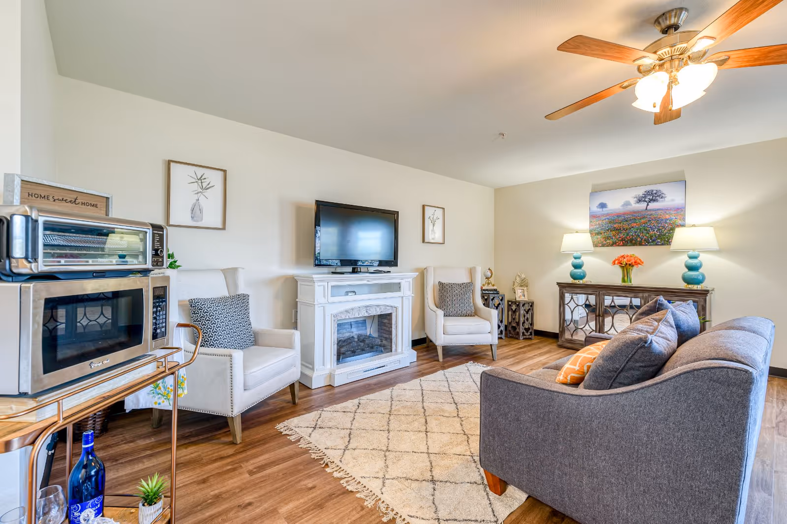 A cozy living room with a gray sofa, two beige armchairs with patterned cushions, a white electric fireplace with a flat-screen TV mounted above it, a wooden sideboard with two blue lamps and a vase of flowers, a ceiling fan with lights, and a small cart holding a microwave and toaster oven. The room has wooden flooring and a light-colored area rug with a diamond pattern.