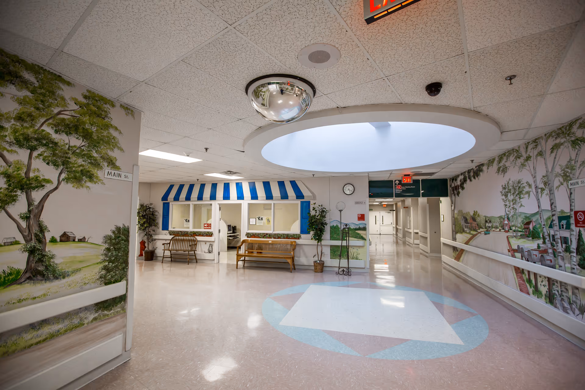 A bright hallway in a senior living facility with murals of trees and houses on the walls. There are benches along the walls, a circular skylight in the ceiling, and a convex security mirror. The floor has a geometric pattern, and there are signs indicating 'Main St.' and an exit sign above a hallway entrance.