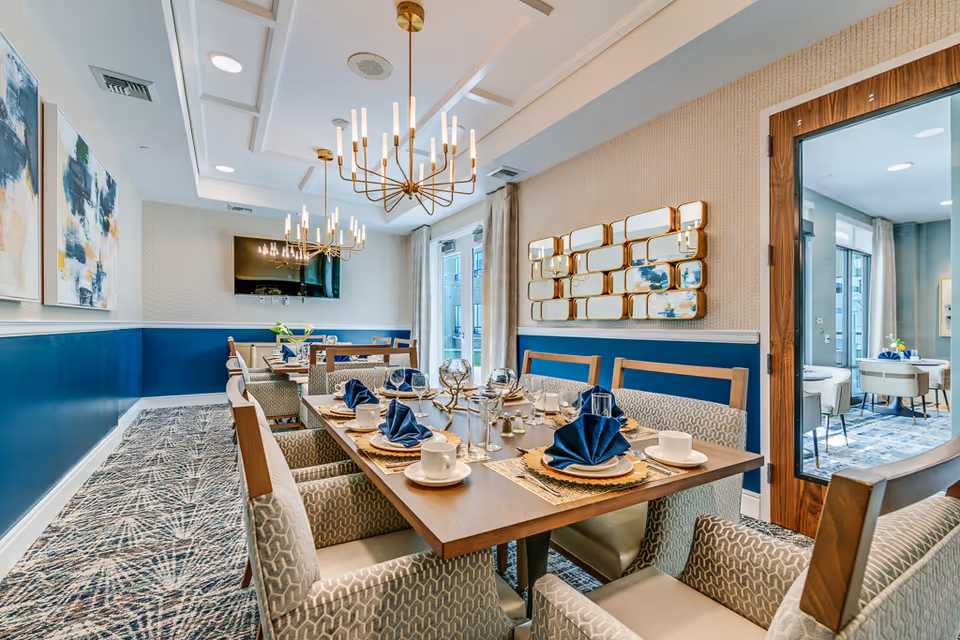 Elegant communal dining room with a long table set with navy napkins, chandeliers, wall art, and upholstered chairs.