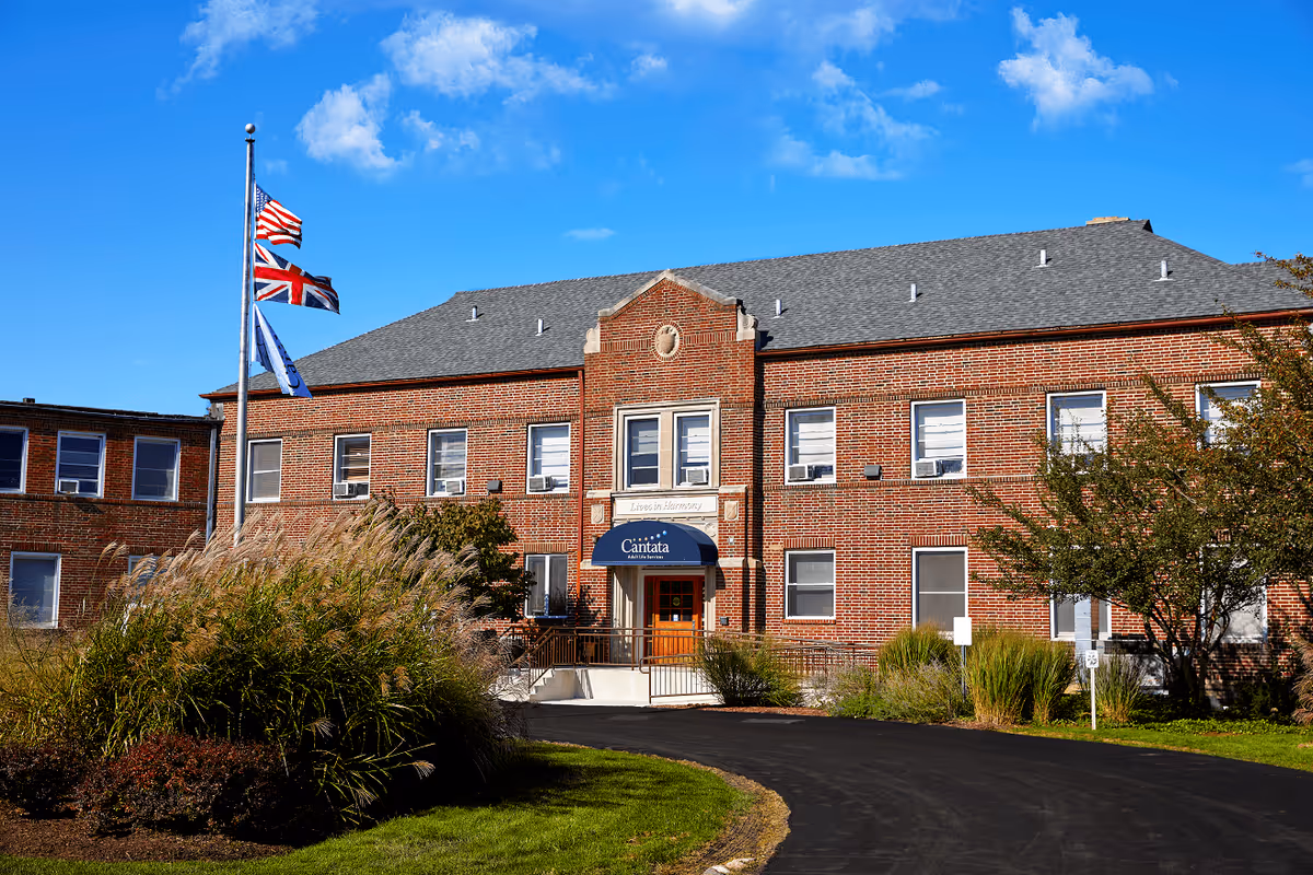 Exterior view of a two-story brick building with multiple windows and a blue awning above the entrance that reads 'Cantata Adult Life Services'. There are three flagpoles in front of the building displaying the American flag, the British flag, and another flag. The building is surrounded by landscaped greenery and a curved paved driveway.
