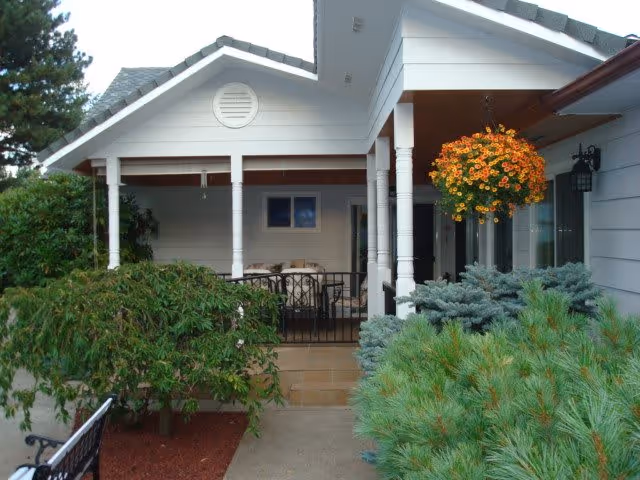 Front porch area of a single-story house with white siding and a gray roof. The porch has white pillars and a hanging basket of orange and yellow flowers. There are green shrubs and bushes surrounding the walkway leading to the porch.